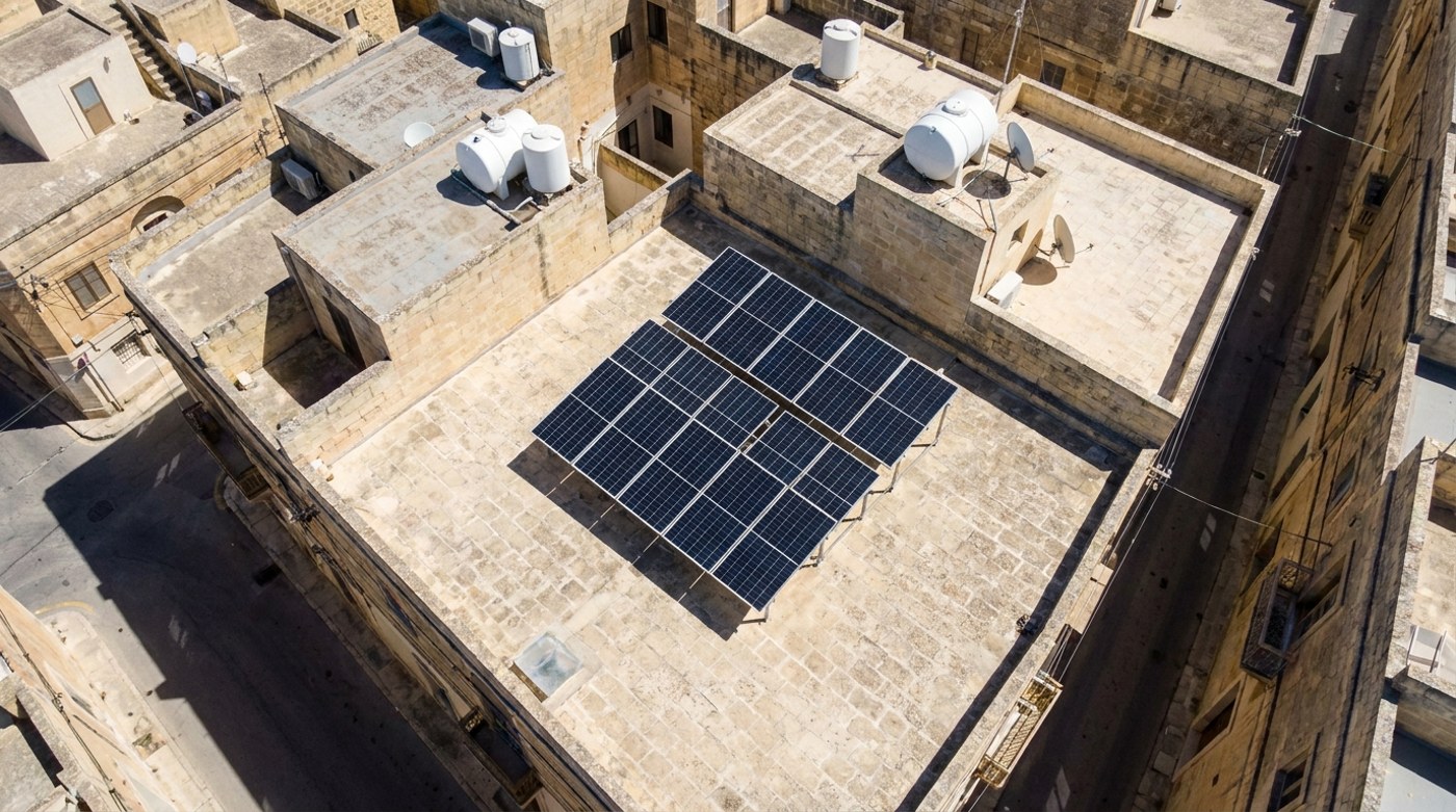 Illustration: drone view looking down on flat limestone rooftops in a Maltese town, one roof with a neat grid of black solar panels and white water tanks on the neighbouring roofs